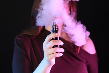 Woman using electronic smoking device in neon lights on black background, closeup
