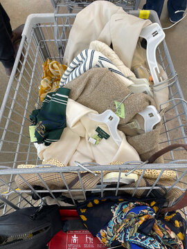 A shopping cart filled with assorted thrift store clothing and accessories. Miami, FL, April 2025.
