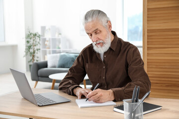 Senior man taking notes while learning online at desk indoors. Self-study