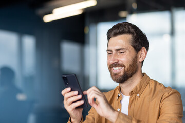 A smiling man in a button-down shirt looks at his phone, likely texting or browsing in a modern...