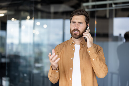 A concerned businessman communicates on his smartphone, gesturing animatedly while looking upwards in an office setting.