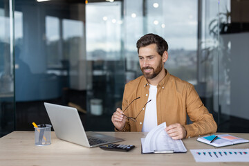 A businessman in a modern office setting reviews documents while using a laptop. He is holding his...