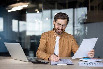 A cheerful businessman with glasses reviews financial documents in his office, using a laptop and...