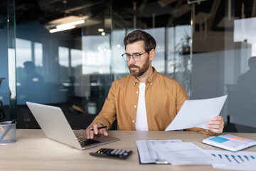 A businessman at his office desk, working on a laptop, holding papers, analyzing data, and wearing...