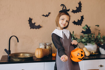 Little girl wearing witch costume holding candies and pumpkin basket celebrating Halloween in...