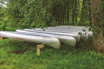 Several metal canoes resting upside down on wooden racks in shaded area near forest, ready for...