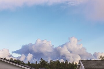 Thunderclouds rising above rooftops and pine trees under blue evening sky in quiet suburban landscape. Sweden.
