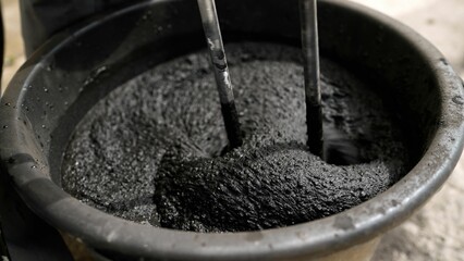 Construction worker mixing cement and other materials in a bucket using two sticks, creating a dark...
