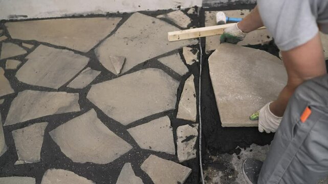 Construction worker carefully placing irregular flagstone on a bed of dark mortar, creating a decorative pathway