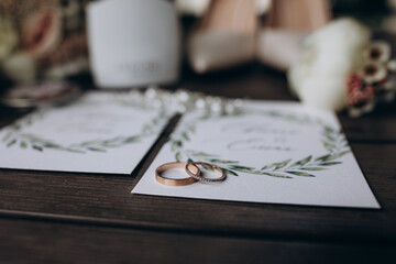 Close-up of two wedding rings placed on elegant invitation cards with greenery design, surrounded...
