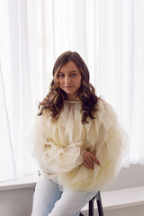 A portrait of a fashionable teenage girl in blouse and sneakers sitting on a chair in a studio on a light wall in a room. light from the window