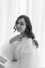 A portrait of a fashionable teenage girl in blouse and sneakers sitting on a chair in a studio on a light wall in a room. light from the window