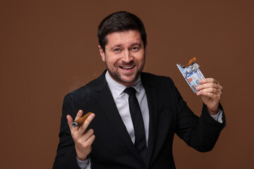 Happy greedy banker with burning dollar banknote smoking cigar on brown background
