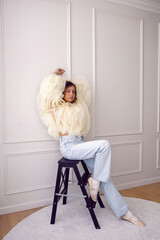 A portrait of a fashionable teenage girl in blouse and sneakers sitting on a chair in a studio on a light wall in a room. light from the window