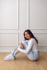 A portrait of a fashionable teenage girl in white clothes and sneakers dancing in a studio on a light wall in a room. light from the window