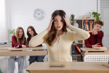 Cyber bullying. Upset teenage girl with smartphone and schoolmates pointing at her in classroom,...