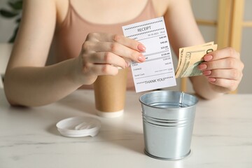Woman with receipt putting tips into metal bucket at white marble table in cafe, closeup