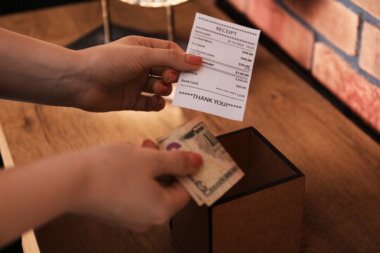 Woman with receipt putting tips into box at wooden table indoors, closeup