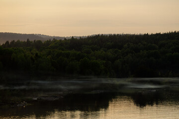 A tranquil scene of mist rising over a forested lake as the soft light of dusk creates a serene...
