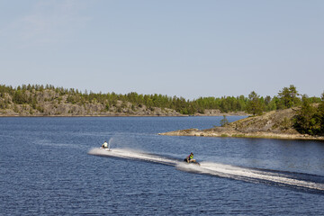 Two people ride jet skis across a clear blue lake surrounded by rocky forested shores on a sunny...