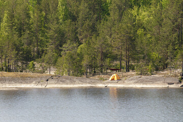 Bright yellow camping tent pitched on rocky shoreline among pine trees, reflecting in calm water...