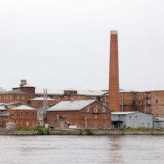 Historic red brick industrial factory complex with tall chimney located along a calm riverside, featuring aged architecture and utility buildings.