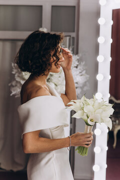 Happy bride in an off-shoulder white gown poses with a bouquet in a stylish, softly lit dressing room. Smiling Bride Holding Flowers in Elegant Dressing Room