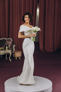 Happy bride in an off-shoulder white gown poses with a bouquet in a stylish, softly lit dressing room. Smiling Bride Holding Flowers in Elegant Dressing Room