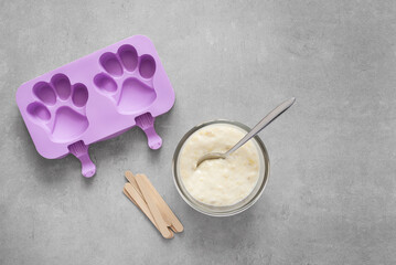 Purple silicone mold in paw shape, creamy mixture in a bowl, and wooden sticks on a gray concrete...