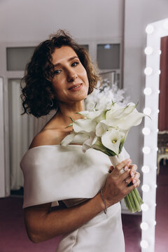 Happy bride in an off-shoulder white gown poses with a bouquet in a stylish, softly lit dressing room. Smiling Bride Holding Flowers in Elegant Dressing Room