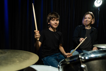 Young caucasian woman teaches a boy to play the drums in the studio on a black background. Music...