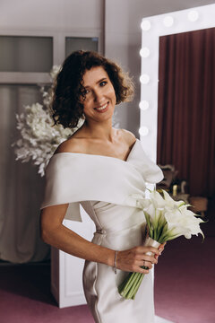 Happy bride in an off-shoulder white gown poses with a bouquet in a stylish, softly lit dressing room. Smiling Bride Holding Flowers in Elegant Dressing Room