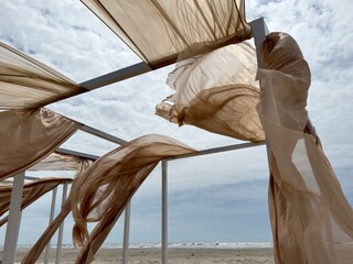 Beige, translucent fabric blows in the wind around a white metal pergola set on a sandy beach under...