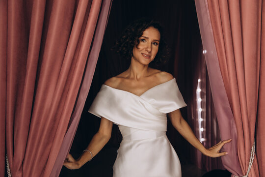 Stylish bride in a fitted off-shoulder gown posing gracefully between parted pink velvet curtains in a dramatic setting. Elegant Bride Framed by Pink Velvet Curtains