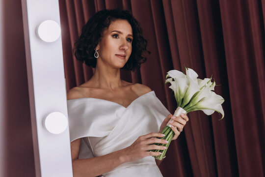 Happy bride in an off-shoulder white gown poses with a bouquet in a stylish, softly lit dressing room. Smiling Bride Holding Flowers in Elegant Dressing Room