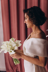Happy bride in an off-shoulder white gown poses with a bouquet in a stylish, softly lit dressing room. Smiling Bride Holding Flowers in Elegant Dressing Room