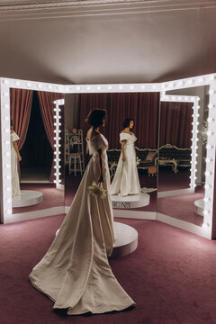 Elegant bride in a long gown holding flowers and looking at her reflection in a triple vanity mirror with dramatic lighting. Bride Admiring Reflection in Illuminated Mirror Setup