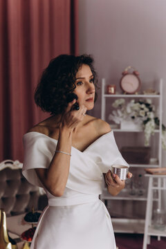 Bride in an off-shoulder gown applies face powder with a brush while getting ready in a softly lit, stylish room. Bride Applying Makeup in Elegant Bridal Suite
