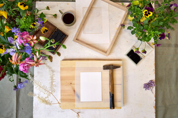 Top view of creative botanical printmaking setup with hammer, flowers, frame, and natural elements...