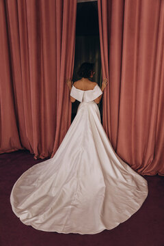 Elegant bride in a long train wedding gown holding flowers while posing gracefully between tall pink velvet curtains. Bride in Flowing Gown Holding Flowers by Velvet Curtains