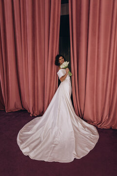 Elegant bride in a long train wedding gown holding flowers while posing gracefully between tall pink velvet curtains. Bride in Flowing Gown Holding Flowers by Velvet Curtains