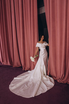 Elegant bride in a long train wedding gown holding flowers while posing gracefully between tall pink velvet curtains. Bride in Flowing Gown Holding Flowers by Velvet Curtains