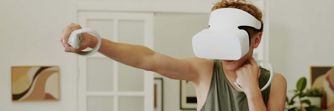 Website banner of Caucasian teenage boy wearing VR headset standing indoors, holding motion controller in outstretched arm, engaging in virtual reality exercise or gaming activity at home - Powered by Adobe