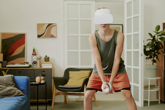 Caucasian teenage boy wearing VR headset exercising with motion controller in modern living room, standing with legs apart and focusing on virtual fitness activity at home