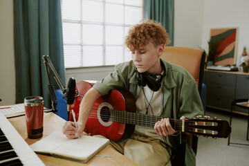 Caucasian teenage boy sitting at desk writing music notes in notebook while playing acoustic guitar, wearing headphones around neck, recording audio with microphone in home studio setup