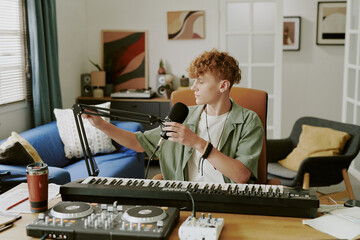 Caucasian teenage boy sitting at desk adjusting microphone while recording music at home, electronic keyboard and DJ controller on table, looking away from camera