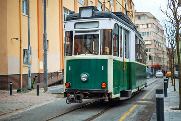 A vintage green tram with white trim travels along tracks on the Kadikoy-Moda line in Istanbul, surrounded by urban buildings and utility poles. A nostalgic mode of public transport in a city setting.