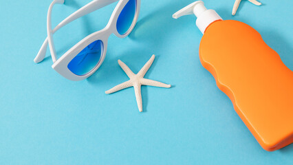 Orange bottle of sunscreen lotion, starfish, sunglasses on blue background. Safe tanning concept on the beach. Banner.