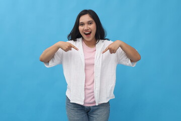 Fototapeta premium Young Asian woman smiling happily looking at the news, pointing down and looking in amazement at the camera, showing an advertisement in the bottom copy space, standing on a blue background.