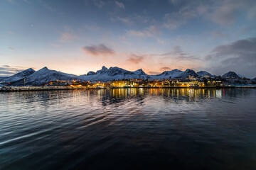 Husøy auf der Insel Senja in Norwegen an einem kalten Winterabend, Sonnenuntergang hinter verschneiten Bergen, isolierte Inselsiedlung in ruhiger Küstenlandschaft und goldener Abendstimmung
